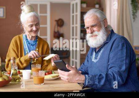 Grandfather having video call on smartphone while having breakfast with his wife at table in the room Stock Photo