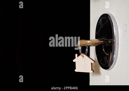 Key with a trinket in the form of a house in the door lock of an open door against the dark background Stock Photo