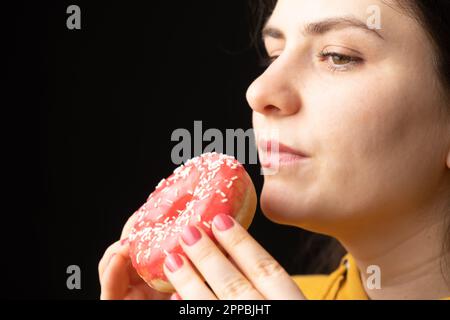 A woman bites a large red donut, a black background, a place for text. Gluttony, overeating and sugar addict. Stock Photo