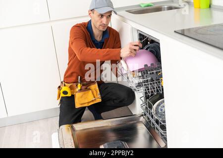 A man or a service worker in special clothes using a screwdriver attaches to the countertop or removes the dishwasher. Stock Photo