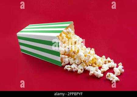 Tasty cheese popcorn falling out of a green striped carton bucket, isolated on red background. Scattering of popcorn grains. Movies, cinema and entert Stock Photo