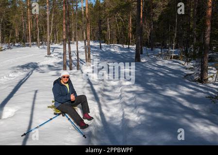 Elderly woman sits in the snow and eats a fruit after walking on frozen snowmobile tracks in the forest. Stock Photo