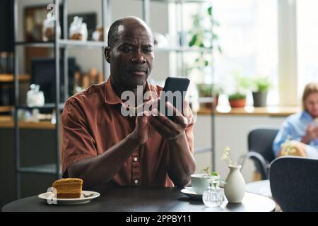 Serious mature man with mobile phone texting or looking through online news while sitting by table in cafe and having tea with cake Stock Photo
