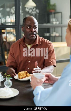 Mature man in brown shirt talking to his wife while both sitting by served table in coffee shop or restaurant and having dessert with tea Stock Photo