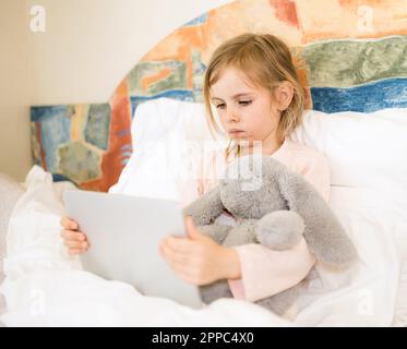 Sick child with red pimples sitting in bed hugging fluffy toy. Little girl looking at tablet. Chickenpox, varicella virus or vesicular rash on child's Stock Photo