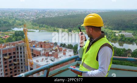 Portrait of Caucasian handsome man builder manager in uniform and helmet looking at digital tablet and talking in walkie-talkie while standing on Stock Photo