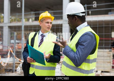 Engineer standing with builder supervising the construction process with house drawings on structure outdoors Stock Photo