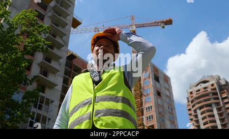 Bottom view of happy handsome young Caucasian man builder standing outdoor taking off yellow helmet on sunny day. Close up. Construction business Stock Photo