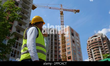 Law angle view of handsome male builder in helmet using walkie-talkie standing at construction site on street. Rear of man constructor speaking in Stock Photo
