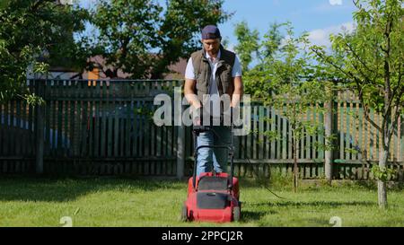 Caucasian handsome guy mowing grass with an electric lawn mower in garden with trees. Young man gardener mows the grass in own courtyard using grass Stock Photo