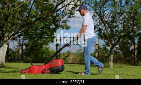 Side view of handsome male worker working in private garden mowing lawn with roller machine. Man walks in the yard and mows green grass with lawn Stock Photo