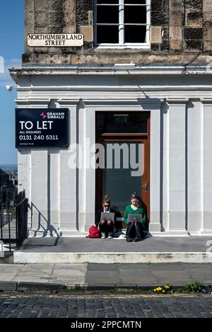 Two young woman in Edinburgh's New Town, sitting in a doorway whilst working on their laptops and enjoying the Spring sunshine. Stock Photo