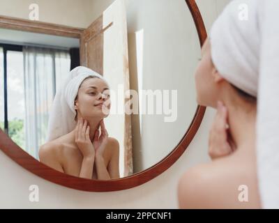 A beauty woman stands in front of a mirror after a shower in a towel on her head looks at her reflection and does a facial massage applies a day cream Stock Photo