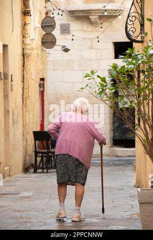 Rabat, Malta - November 13, 2022: Super senior woman walking with a cane in the pedestrian street, from behind Stock Photo