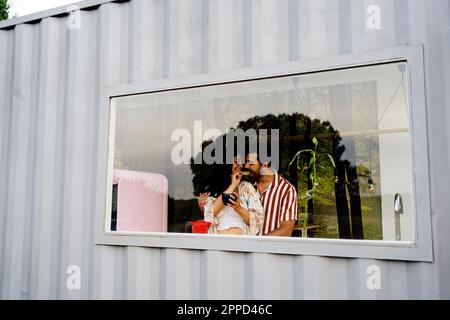 Woman feeding boyfriend in kitchen seen through window of container home Stock Photo