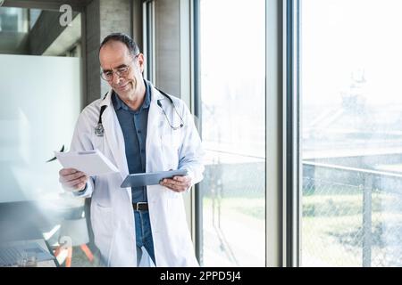 Smiling mature doctor reading prescription and holding tablet PC by window Stock Photo