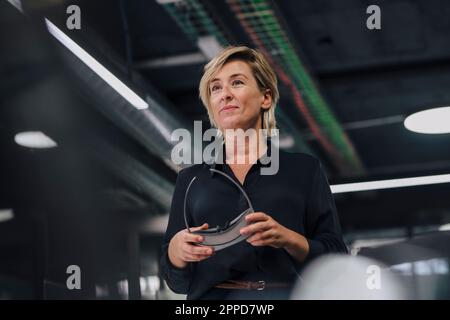 Contemplative smiling businesswoman standing with VR glasses in office Stock Photo