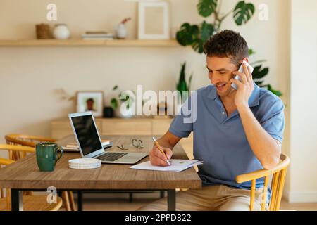 Smiling freelancer talking on mobile phone and taking notes on paper at table Stock Photo