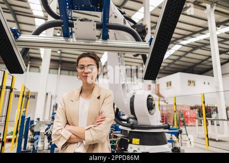 Young engineer standing with arms crossed in front of robotic arm Stock Photo