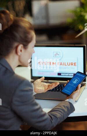Seen from behind modern woman with laptop and smartphone experiencing computer malfunction and having voice bot artificial intelligence conversation i Stock Photo