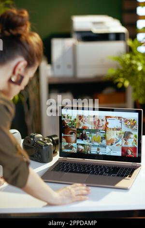 Seen from behind female photographer with laptop and photo camera viewing photos in modern office. Stock Photo