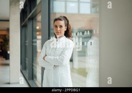 Confident woman in lab coat standing with crossed arms near window Stock Photo