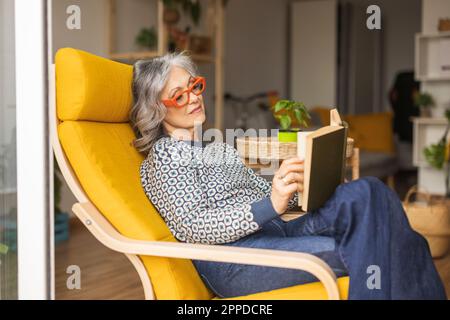 Mature woman reading book sitting on armchair Stock Photo
