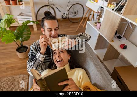 Smiling man eating snack with girlfriend reading book lying on sofa at home Stock Photo