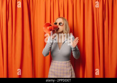 Woman making announcement through megaphone in front of orange curtain Stock Photo