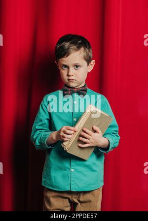 Boy holding book standing in front of red curtain Stock Photo