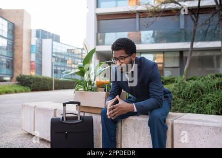 Depressed businessman with suitcase and his belongings sitting outside office building Stock Photo