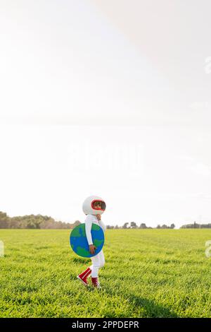 Girl wearing astronaut costume holding earth cut out standing on grass Stock Photo