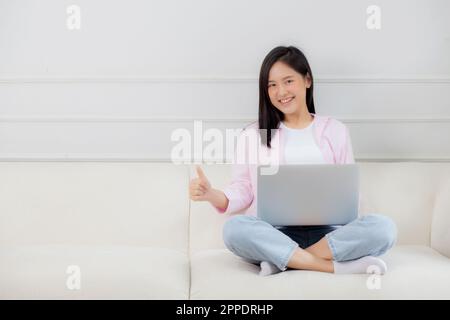 Young asian business woman working from home with laptop computer and thumbs up sitting on sofa with success on sofa in living room at home, freelance Stock Photo