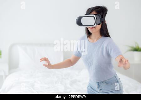 Young asian woman wearing vr headset playing game with excited in the bedroom at home, female using virtual reality or metaverse innovation for simula Stock Photo