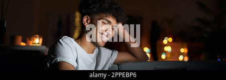 Smiling young guy watching movie online using laptop and eating popcorn while sitting on a comfortable sofa in dark room at home Stock Photo
