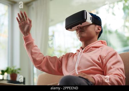 Young man exploring metaverse when sitting on couch at home in VR headset Stock Photo