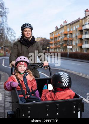 Young mother cycling with two children in cargo bicycle Stock Photo