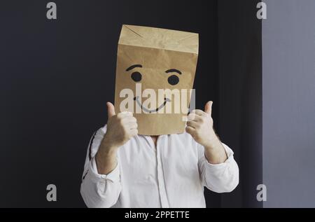 A man in a white shirt with a paper bag on his head, with a smiley face drawn, stands against the wall, overwhelmed with feelings, shows a hand gestur Stock Photo