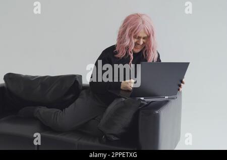 Portrait of a man in a pink wig, sitting on a sofa, typing on a laptop. White background. Stock Photo