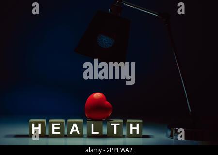 Wooden cubes with the inscription HEALTH on a table in total darkness and the glow of a table lamp illuminating a red heart Stock Photo