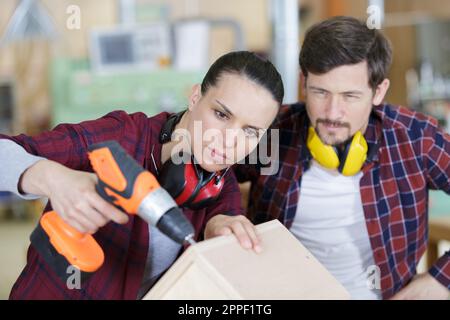 happy female carpenter drilling wood while colleague working in background Stock Photo