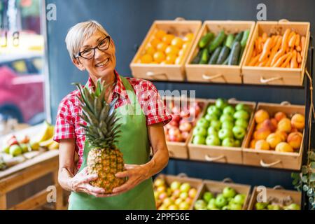 Mature woman works in fruits and vegetables shop. She is holding pineapple. Stock Photo