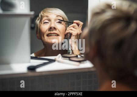 Mature woman is applying mascara in the bathroom. Stock Photo