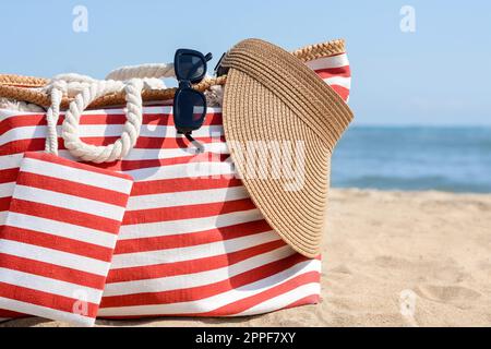 Stylish striped bag with visor cap, sunglasses and blanket on sandy beach  near sea Stock Photo