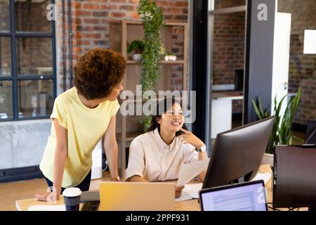 Two happy diverse female creative colleagues looking at computer and discussing at desk in office Stock Photo