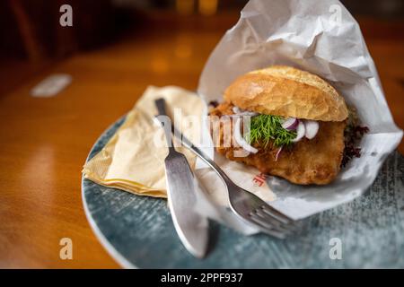 Close up of a fried chicken sandwich wrapped in paper on a plate with fork and knife on a wooden table in a fast food restaurant. Stock Photo
