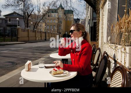 Portrait beautiful tourist woman drinks cup coffee and eats cake, sits in street cafe in city center. Elegant girl wears red blazer jacket, white turt Stock Photo