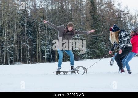 Three girls pulling young man standing on sled in snowy landscape, Bavaria, Germany Stock Photo