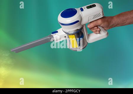 The male hand holds hold a modern cordless battery-powered vacuum cleaner over abstract blurred background. Space. Household cleanliness and hygiene. Stock Photo