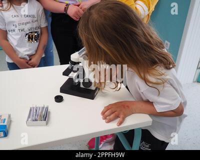 Sremska Mitrovica, Serbia, May 15, 2021. A girl of 7 years old in a white T-shirt looks with one eye through a microscope. Biology lesson. The process of teaching children. School lessons Stock Photo